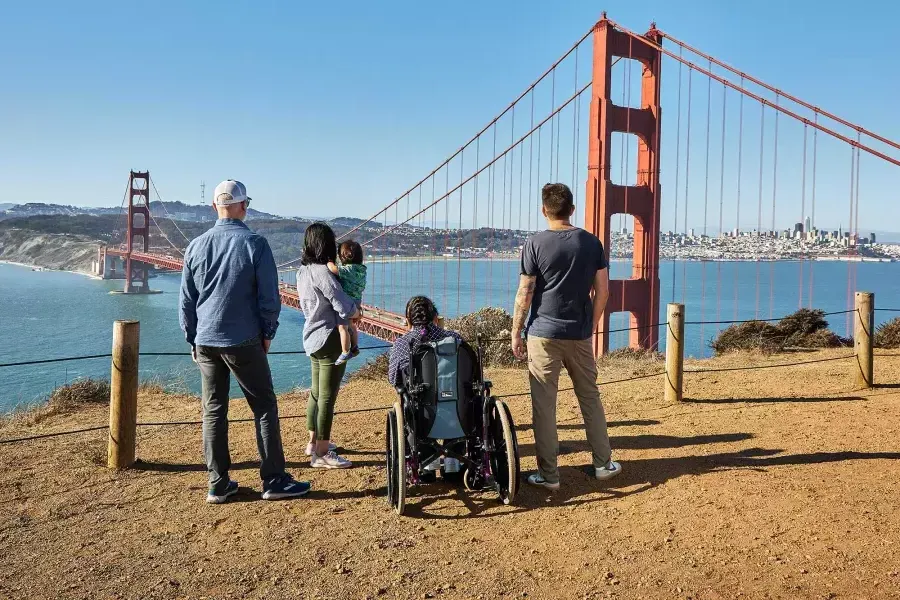 Se ve a un grupo de personas, incluida una persona en silla de ruedas, desde atrás mientras observan el Golden Gate Bridge desde Marin Headlands.